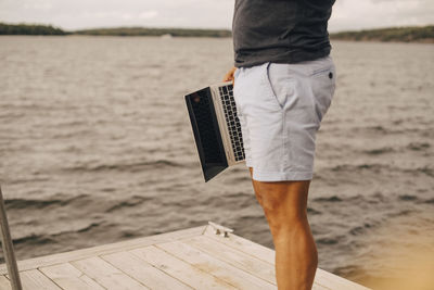 Midsection of man holding laptop while standing on jetty over lake