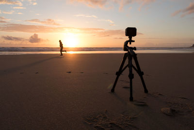 Tripod camera at beach against sky during sunset