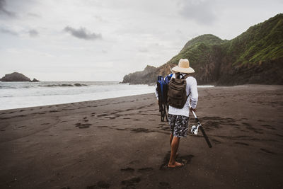 Full body back view of anonymous man with fishing rod with rucksack strolling on sandy shore near sea in nature