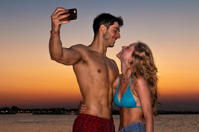 Young man photographing woman using smart phone against sunset sky