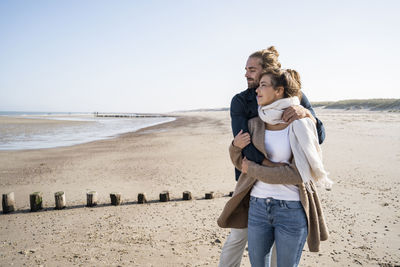 Young couple with eyes closed embracing while standing at beach against clear sky