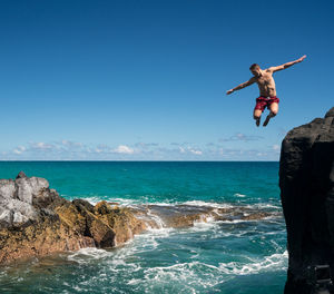 Young man jumping in sea from rock against sky