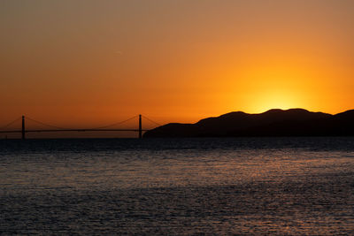 Silhouette bridge over sea against orange sky