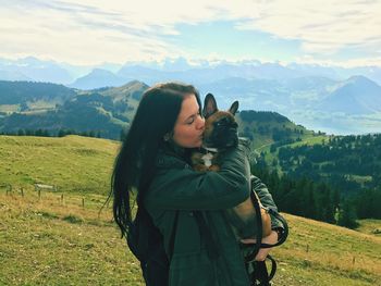 Young woman carrying dog while standing on mountain against cloudy sky