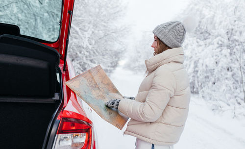 Side view of woman holding map while standing on snow covered land during winter