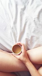 Low section of woman holding coffee cup on bed