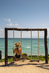 Rear view of woman sitting on swing by sea against sky