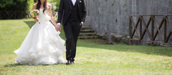 Low section of newlywed couple holding hands and walking on grassy field