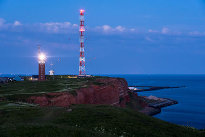 Lighthouse by sea against illuminated buildings against sky