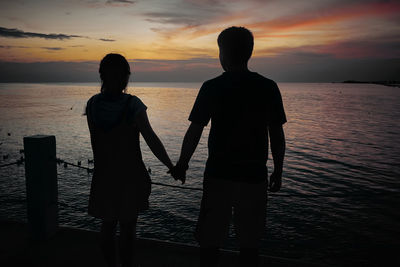 Rear view of silhouette couple holding hands while standing at beach during sunset