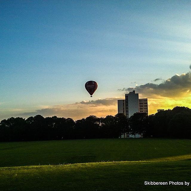 architecture, building exterior, built structure, sky, sport, sunset, tree, hot air balloon, grass, landscape, city, transportation, field, clear sky, flying, modern, mid-air, outdoors, tower, skyscraper