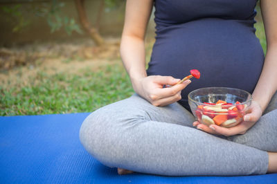 Low section of pregnant woman holding bowl with fruits while sitting on exercise mat