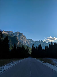 Empty road along trees and mountains against blue sky