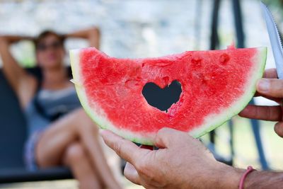 Cropped hand of man holding watermelon while woman relaxing on chair in background