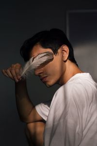 Young man holding feather over eyes while standing against wall