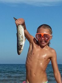 Portrait of happy shirtless girl holding dead fish by sea against sky