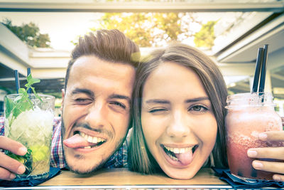 Portrait of cheerful young couple sticking out tongue while having drinks at table