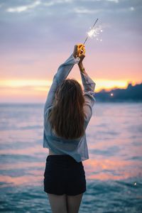 Woman with lit sparkler standing at beach during sunset