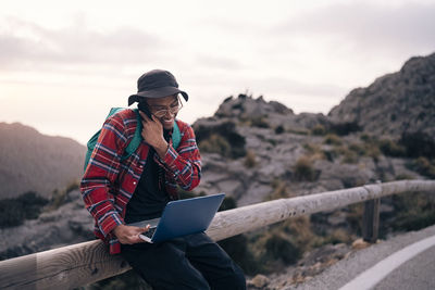 Smiling male explorer talking on smart phone while sitting with laptop on railing