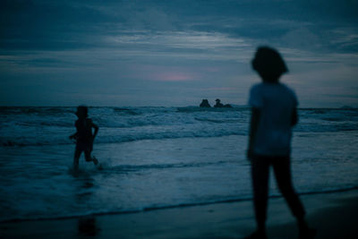 Silhouette siblings at beach against sky during dusk