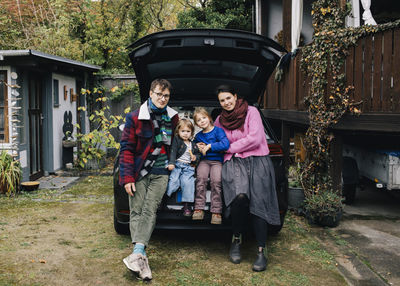 Smiling family sitting together in car trunk outside house