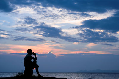 Silhouette man sitting at beach against cloudy sky during sunset