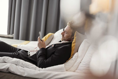 Side view of businessman listening music while lying on bed at hotel room