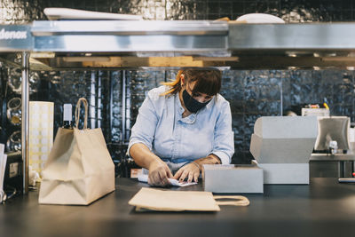 Female chef holding paper by take out containers at restaurant kitchen counter during covid-19