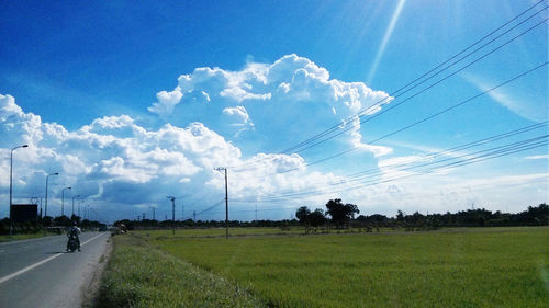 Road by electricity pylons on grassy field against sky