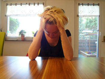 Depressed woman with hand in hair sitting at wooden table in brightly lit room