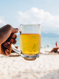 Close-up of hand holding beer glass on beach against sky