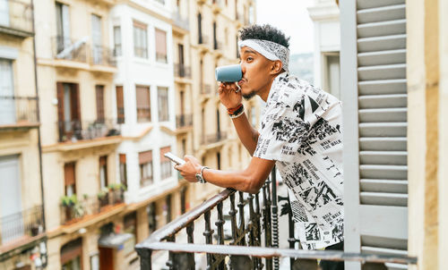 Side view of cheerful ethnic male standing on balcony and browsing smartphone while enjoying morning coffee and looking away