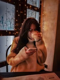 Young woman using smart phone while drinking drink at home