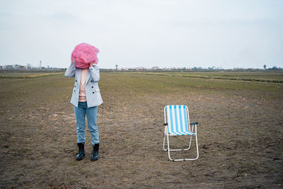 Young woman covering face with tulle net by chair on drought agricultural field