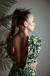 Young woman standing with her back in green floral dress looks to side on gray