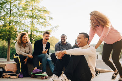 Smiling woman pushing man on skateboard while happy friends sitting at park