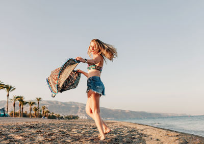 Full length of woman holding scarf while standing at beach against sky