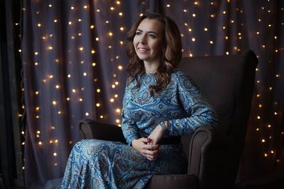 Smiling woman sitting on chair against illuminated lighting equipment