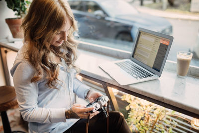 High angle view of young woman holding camera while sitting with laptop at cafe