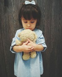 Cute baby girl holding toy while standing against wooden wall