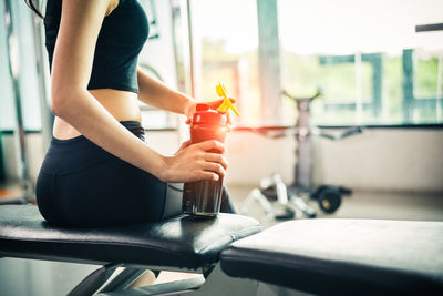 Midsection of young woman holding bottle while sitting on seat in gym