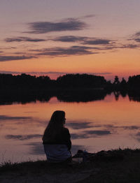 Woman sitting by lake against orange sky