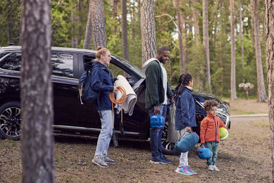 Multi-ethnic family standing with camping gear by car amidst trees