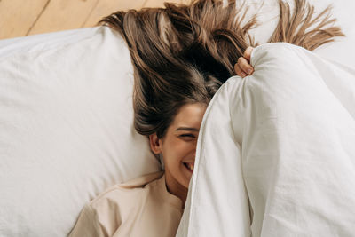 Young cheerful playful brunette woman with long hair hides her face under the covers laughing.