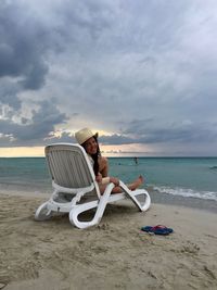 Portrait of young woman relaxing on lounge chair at sea shore against cloudy sky