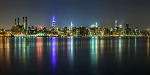 Illuminated cityscape by sea with reflection against sky during night