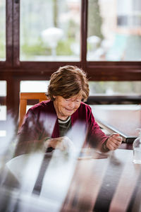 High angle of senior woman in glasses sitting at table and browsing social media on smartphone at home