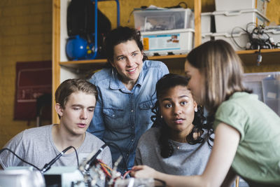 Multi-ethnic teenage students discussing with mature female teacher over science project in classroom at high school