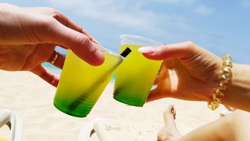 Cropped hands of couple toasting drinks at beach against sky
