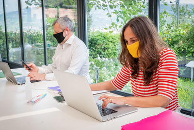 Mature man and woman wearing mask working from home sitting by window